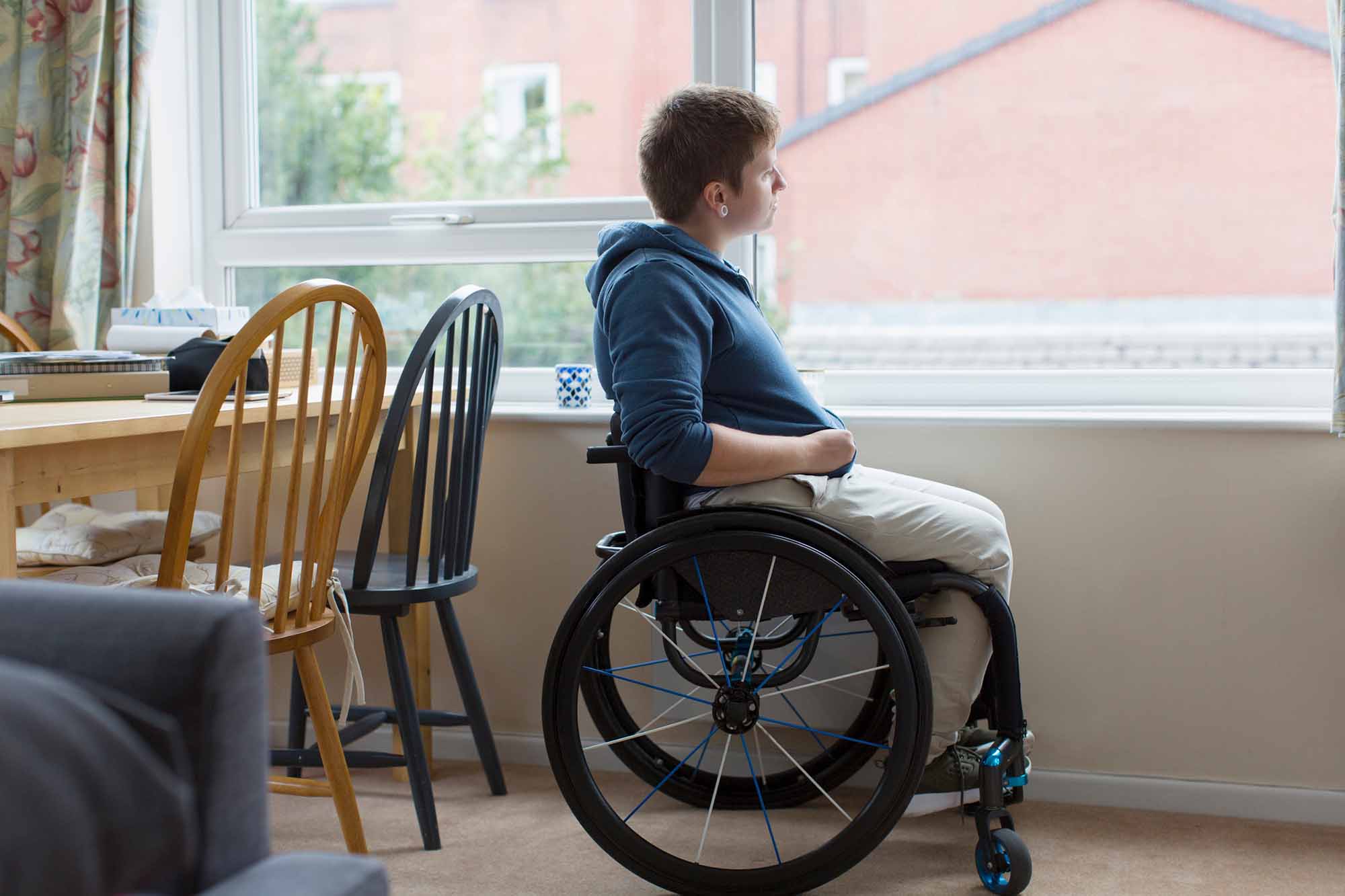 En ung vuxen person som sitter i rullstol i hemmiljö och tittar ut genom ett fönster