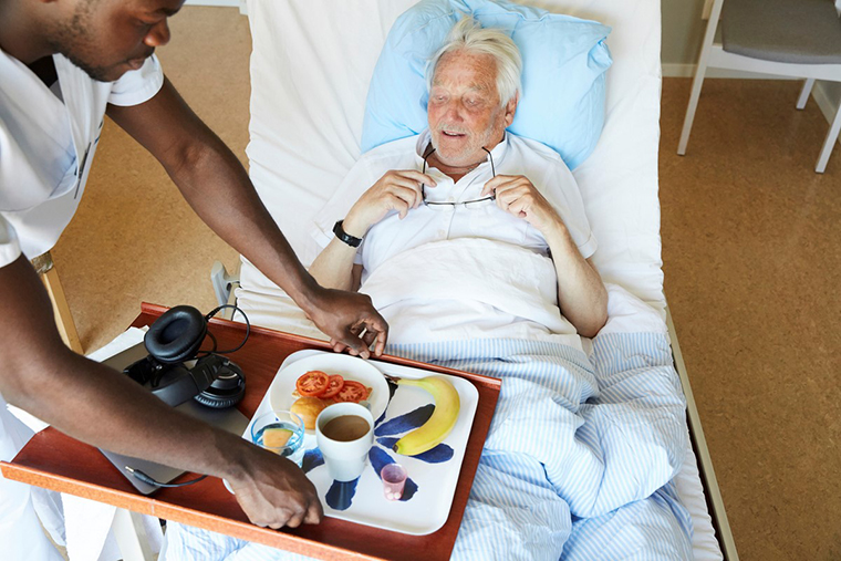 vårdpersonal serverar matbricka till äldre man i sjukhussäng